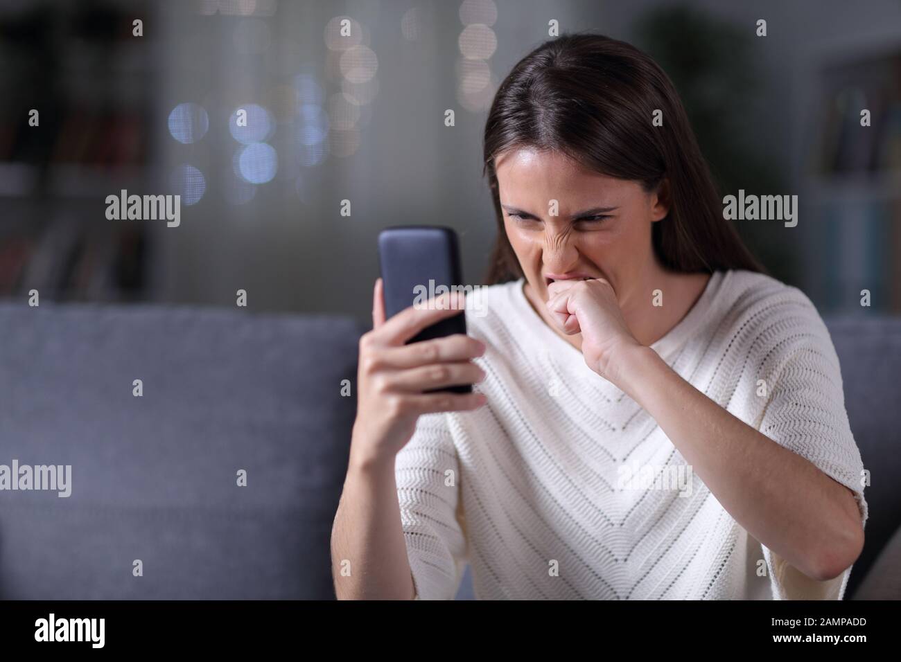 Ragazza arrabbiata che controlla il contenuto del telefono cellulare seduto su un divano nella notte a casa Foto Stock