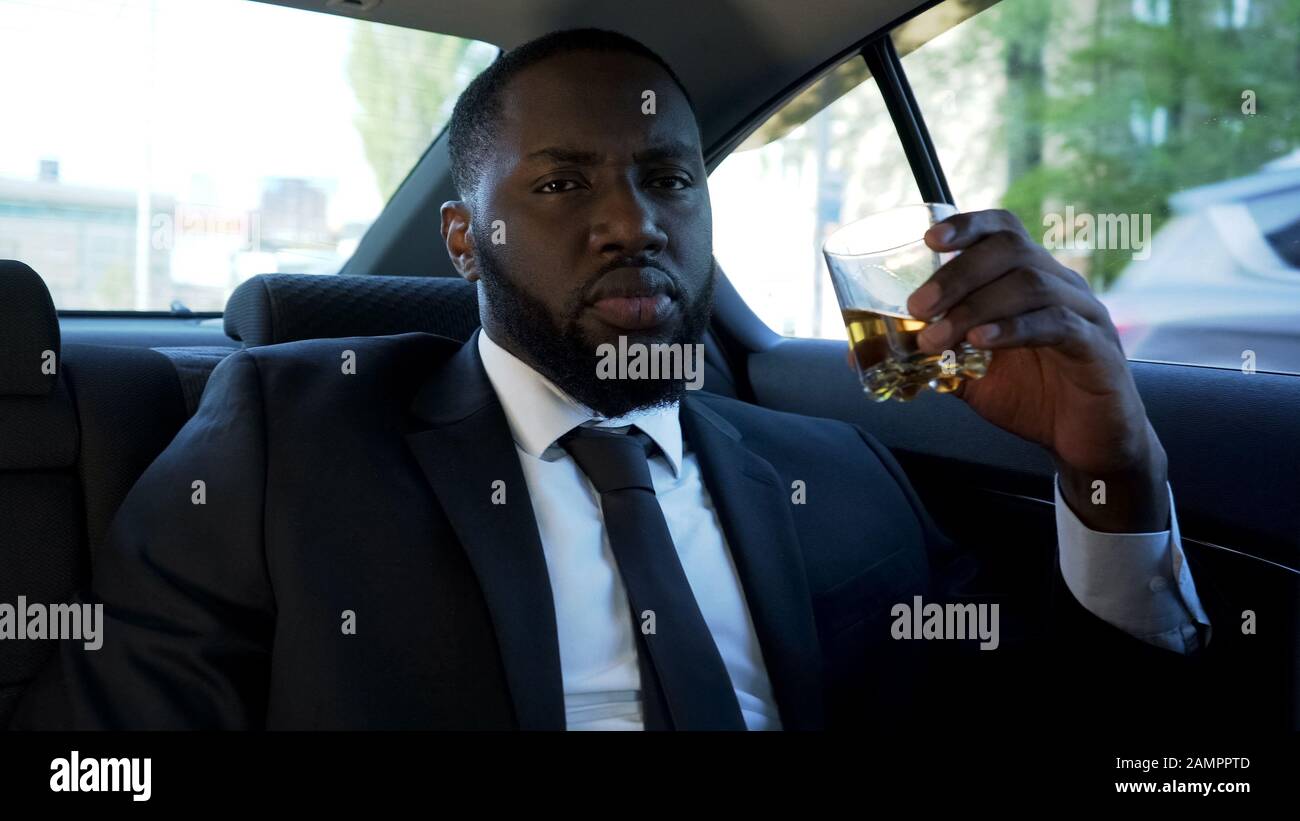 Un uomo d'affari nero di successo bevendo alcool d'elite in automobile, boss mafioso, gangster Foto Stock