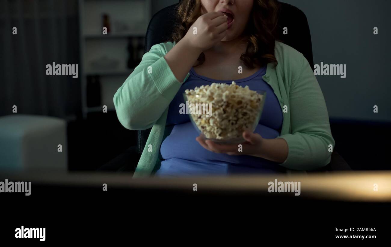 Chubby donna prendere una manciata di popcorn e mangiare di fronte alla TV, pigrizia Foto Stock