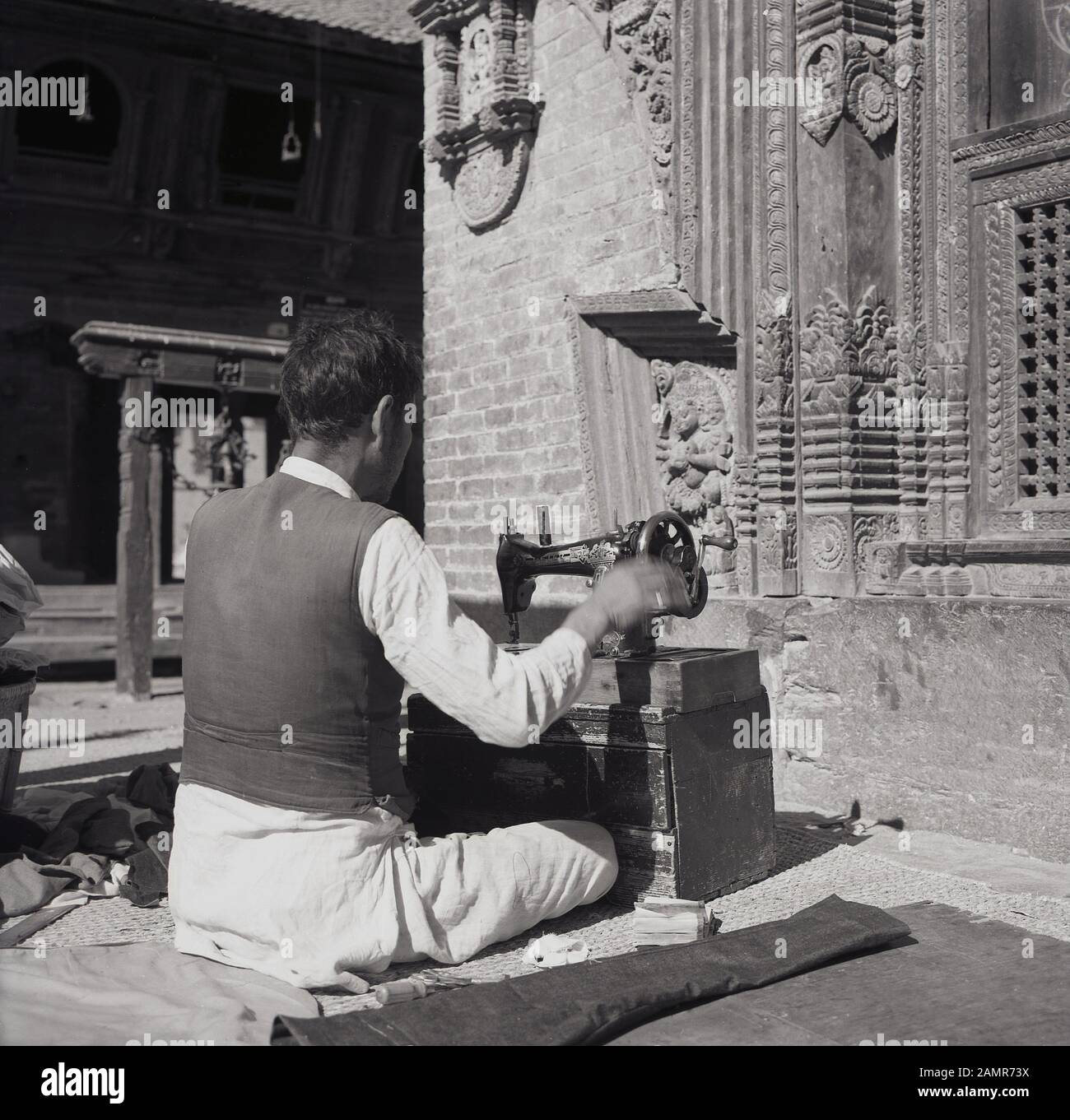 Degli anni Cinquanta, historcial, Nepalese uomo seduto sul terreno al di fuori di un Santuario con una mano-avviato macchina da cucire, Kathmandu, Nepal. In questa epoca, sartoria e riparazione dei vestiti era un importante attività di strada in questo povero mountaineous regione. Foto Stock