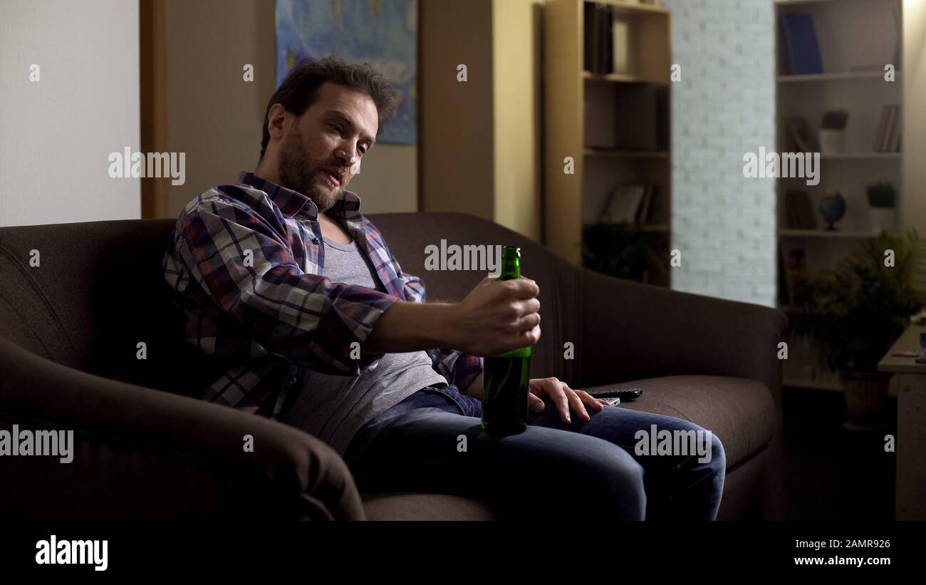 Maschio ubriaco seduto sul divano e parlando con una bottiglia di birra, dipendenza dall'alcol Foto Stock