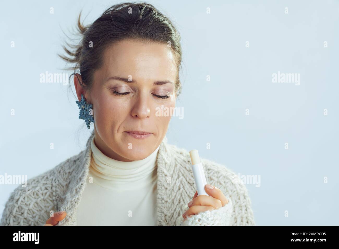 elegante massaia di mezza età in pullover roll collo e cardigan utilizzando labbro balsamo come inverno labbro cura isolato su sfondo azzurro inverno. Foto Stock