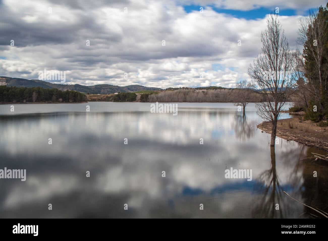 Paesaggio di alberi in acque calme lago con nuvole bianche cielo riflesso e montagne sullo sfondo Foto Stock
