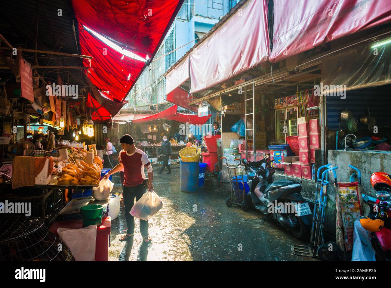 Khlong Toei mercato a Bangkok, Thailandia con bancarelle di cibo sotto un tetto rosso con un uomo che tiene borse di plastica e l'acquisto presso un fornitore in una strada di luce del sole Foto Stock