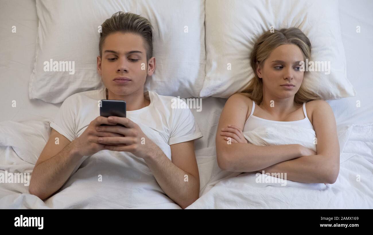 Donna arrabbiata che guarda il ragazzo usando il cellulare, ignorandola a letto, dipendenza Foto Stock