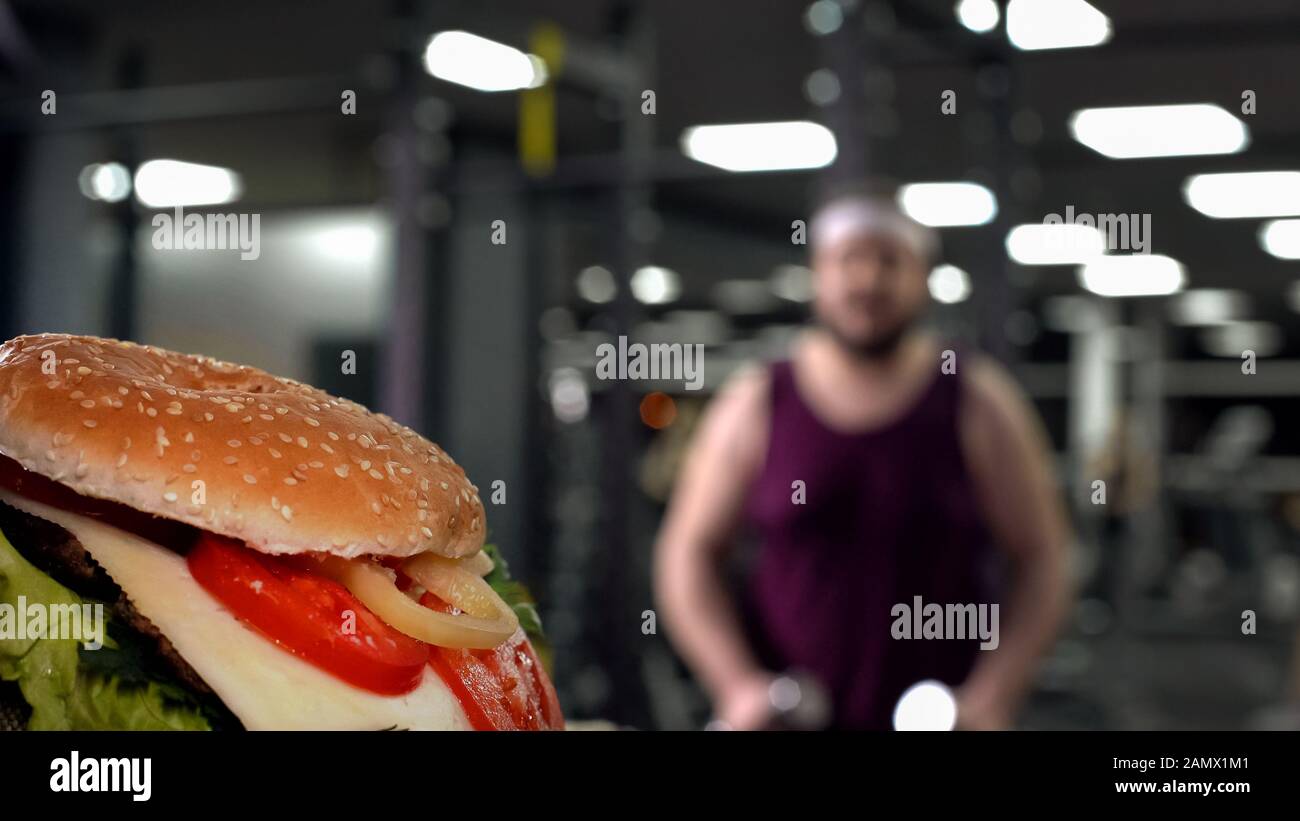 Closeup di hamburger gustoso, uomo chubby con dumbbells sullo sfondo, motivazione Foto Stock