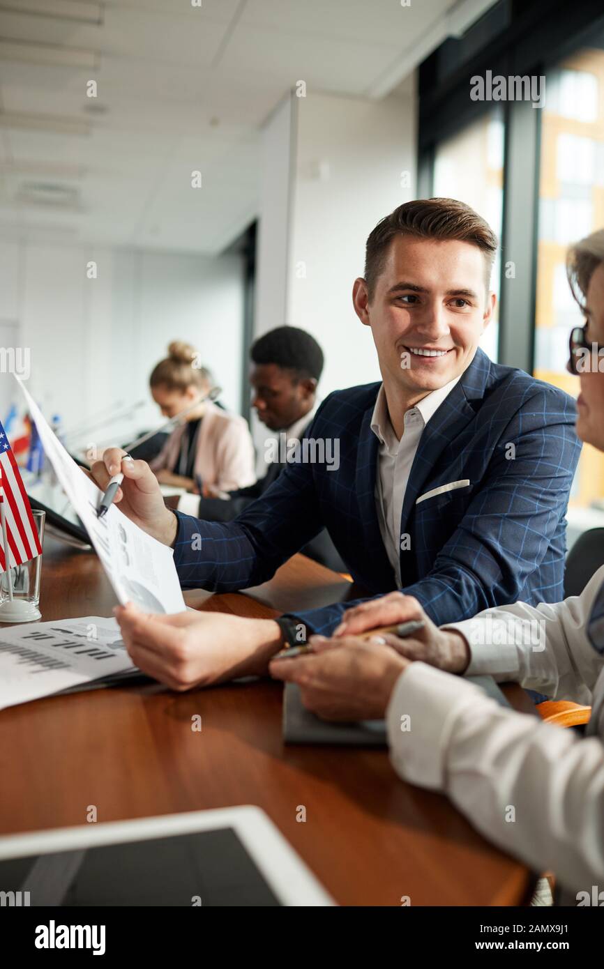 Giovane imprenditore sorridente puntando al documento e di discuterne con il suo partner al tavolo durante la conferenza Foto Stock