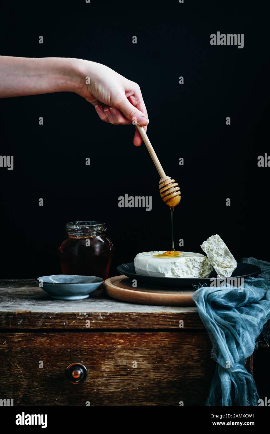 Cottage formaggio con miele su un tavolo rustico su uno sfondo scuro. Mano della donna che tiene un bilanciere di miele e miele che sgocciola da esso Foto Stock