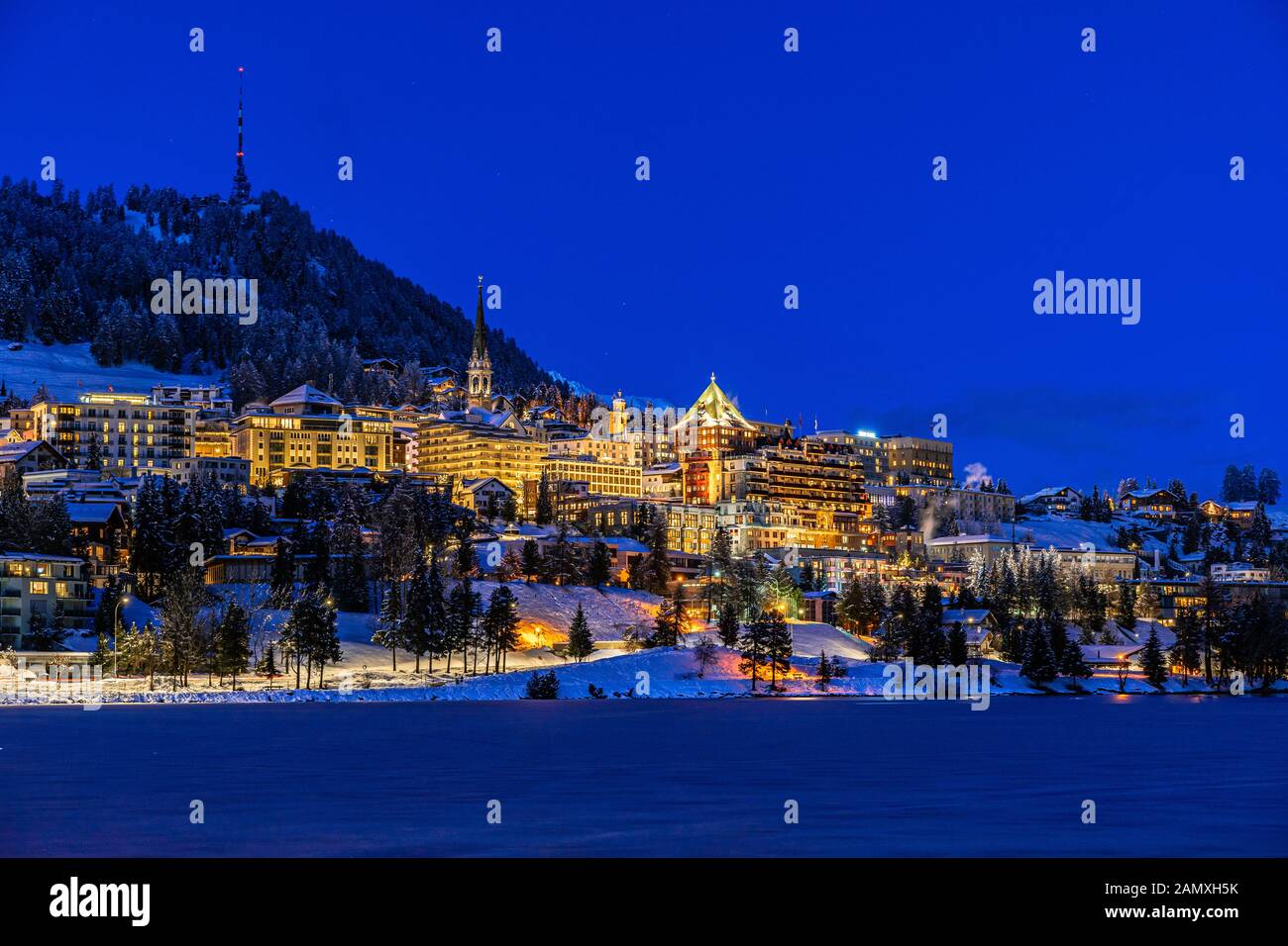 Vista delle belle luci notturne della città di St. Moritz in Svizzera di notte in inverno, con riflessione dal lago e montagne di neve in backgrouind Foto Stock