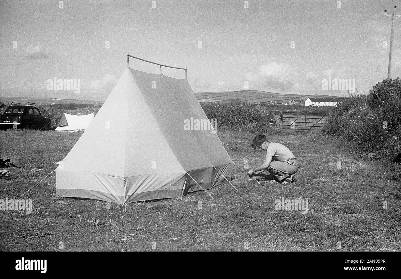 Degli anni Cinquanta, storico, camping, un giovane uomo in un campo tieing nel terreno delle cordicelle della tela di una tenda che ha appena presentato, Inghilterra, Regno Unito. Foto Stock