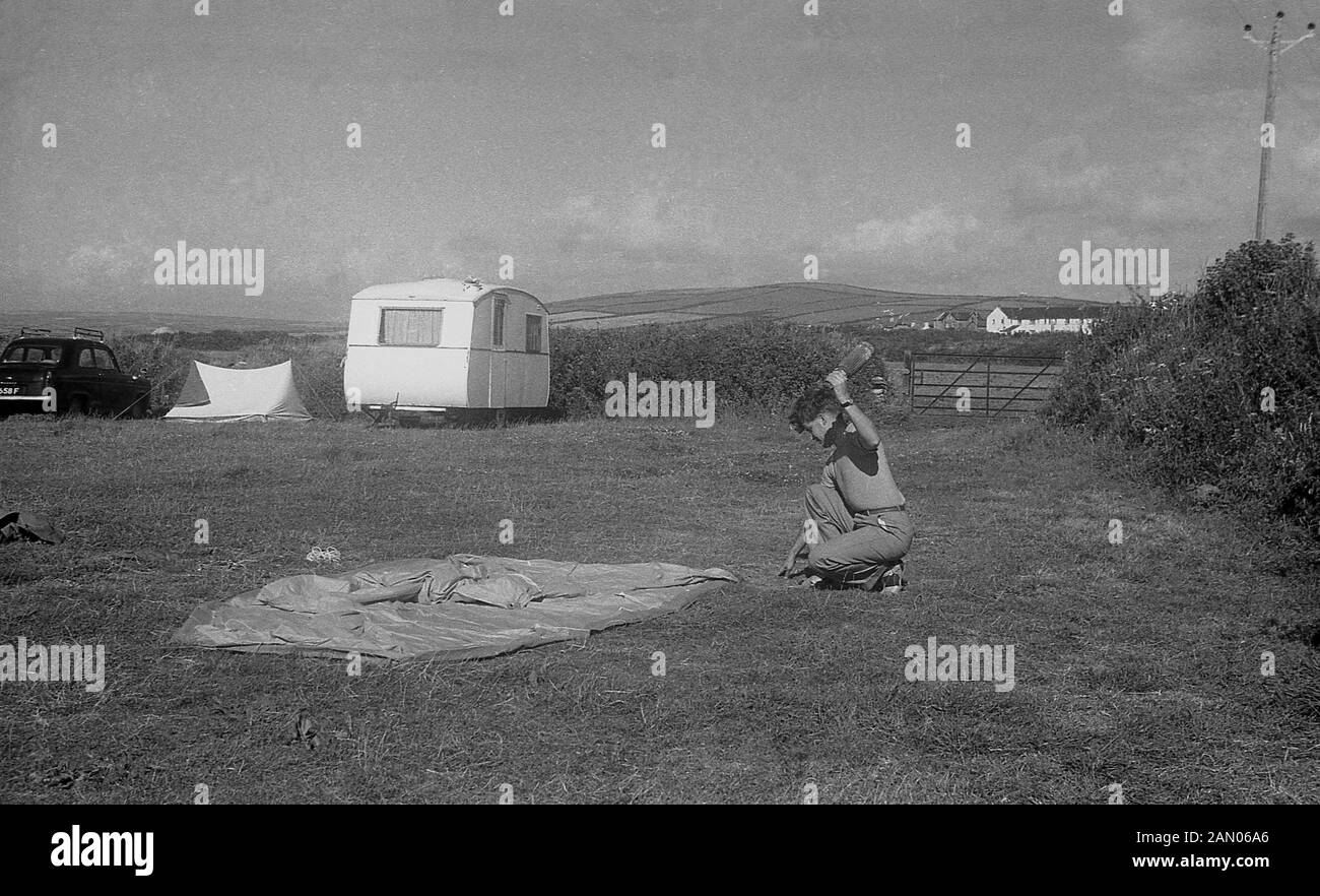 Degli anni Cinquanta, storico, camping, un giovane uomo in un campo in ginocchio sull'erba attorno a mettere una tenda di tela. Foto Stock
