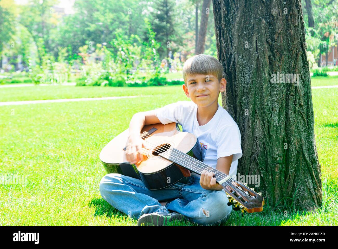 Un ragazzo con una chitarra si siede sotto un albero, canta canzoni e gode di natura. Foto Stock