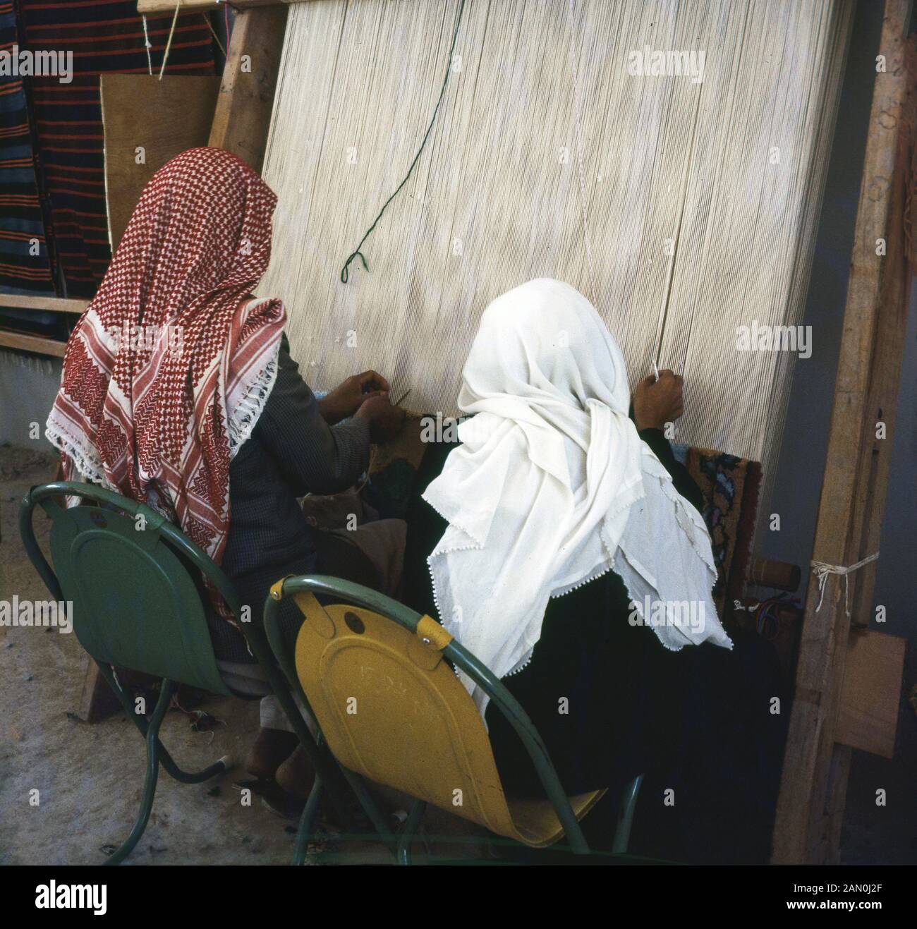 Anni sessanta, storico, due maschi arabi seduti su sedie al di fuori, indossando la Keffiyeh, il tradizionale copricapo arabo, lavorando a mano in corrispondenza di una cornice di legno telaio di tessitura di un dispositivo usato per la tessitura stoffa, Arabia Saudita, Foto Stock