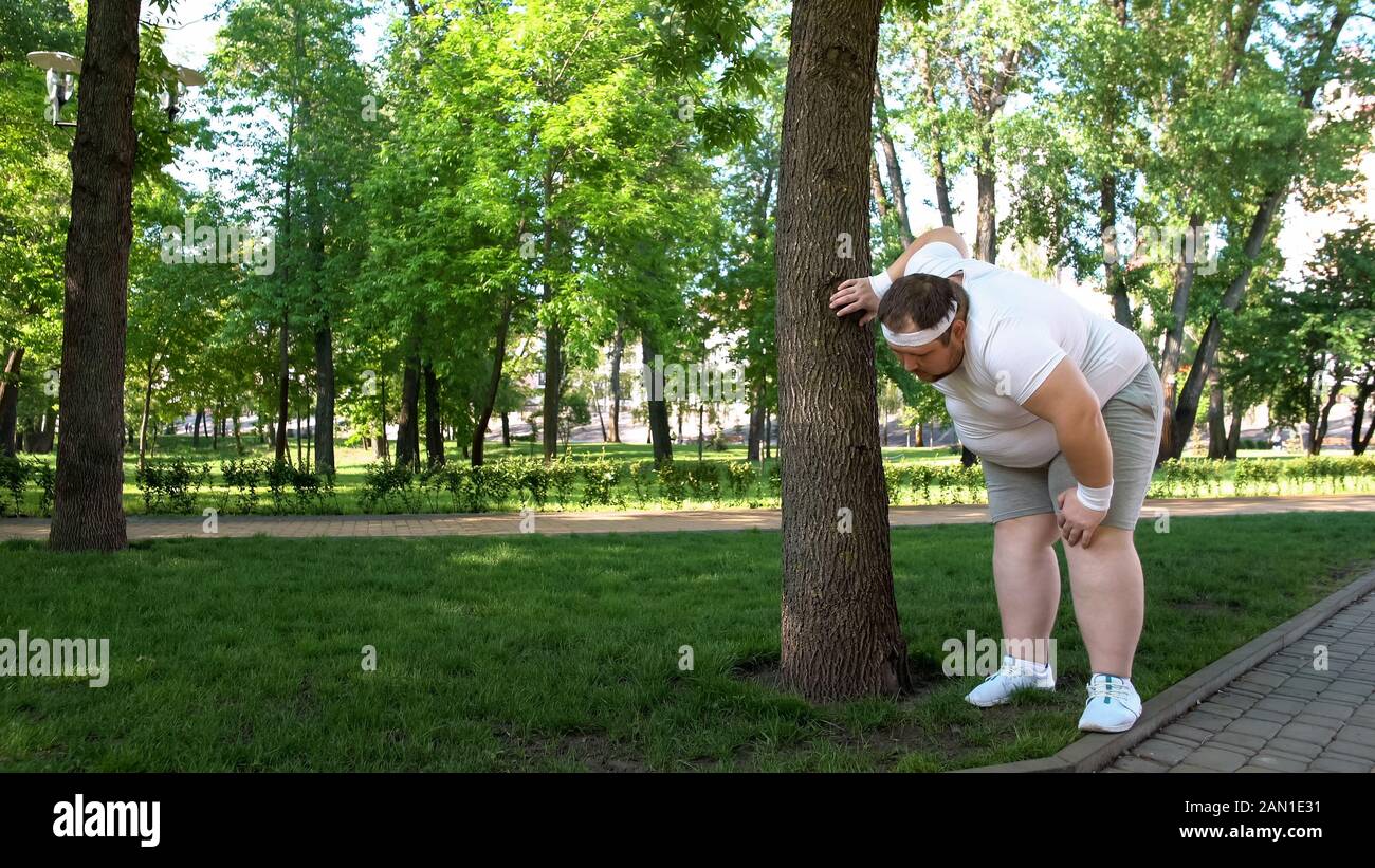 FAT man stanco dopo aver eseguito nel parco, appoggiato su un albero, faticoso workouts all'aperto Foto Stock