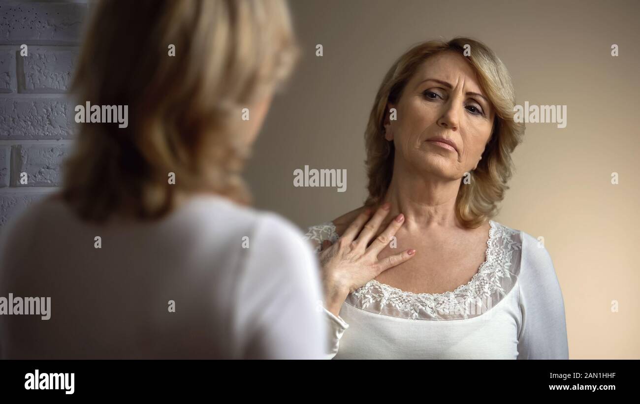 Donna senior infelice che guarda nello specchio e tocca la zona del decollito, le rughe Foto Stock