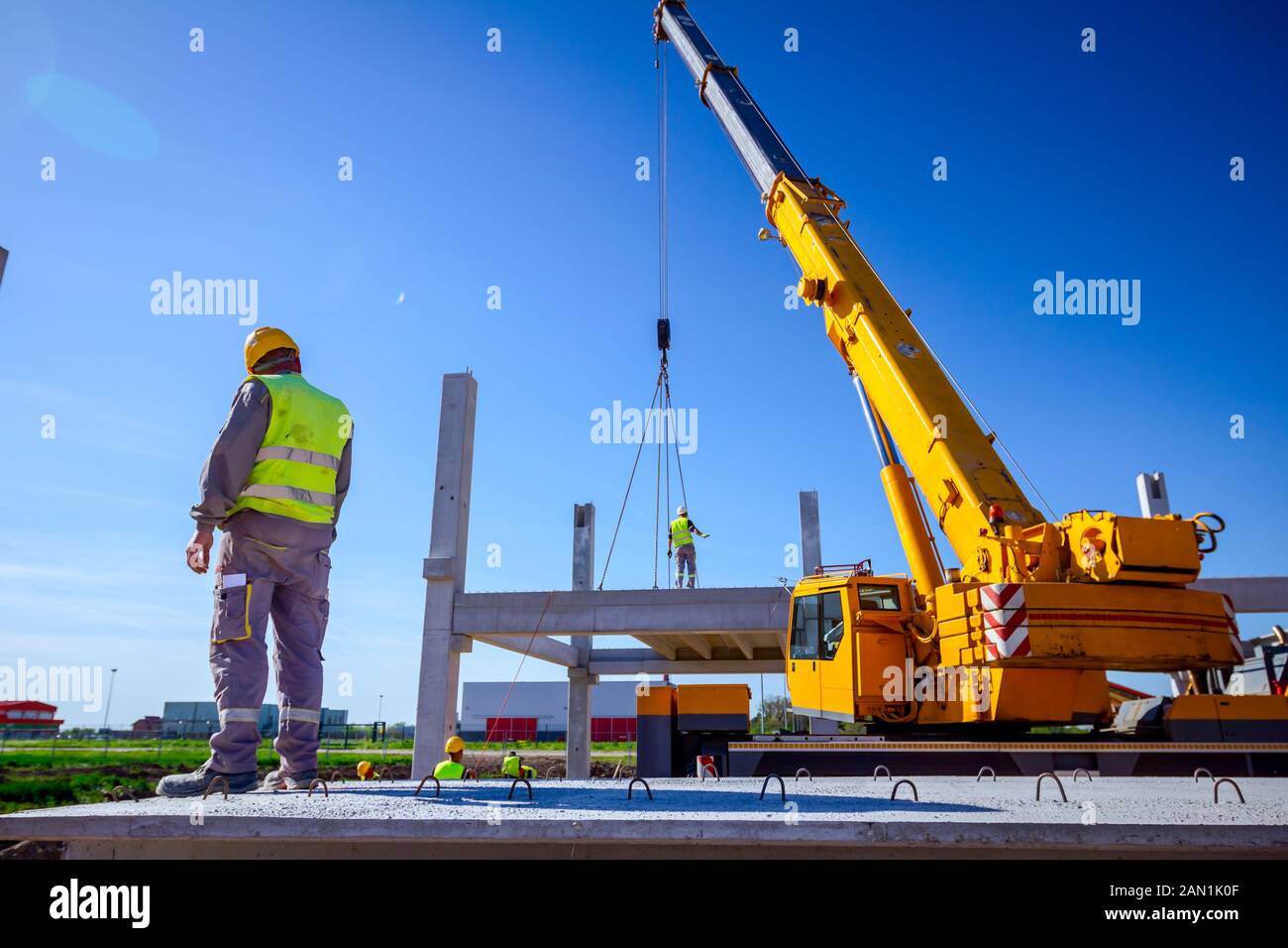 Vista da dietro il lavoratore edile con giubbotto di sicurezza e casco giallo di supervisionare gru mobile per gestire un travetto in cemento per il montaggio enorme costrut Foto Stock