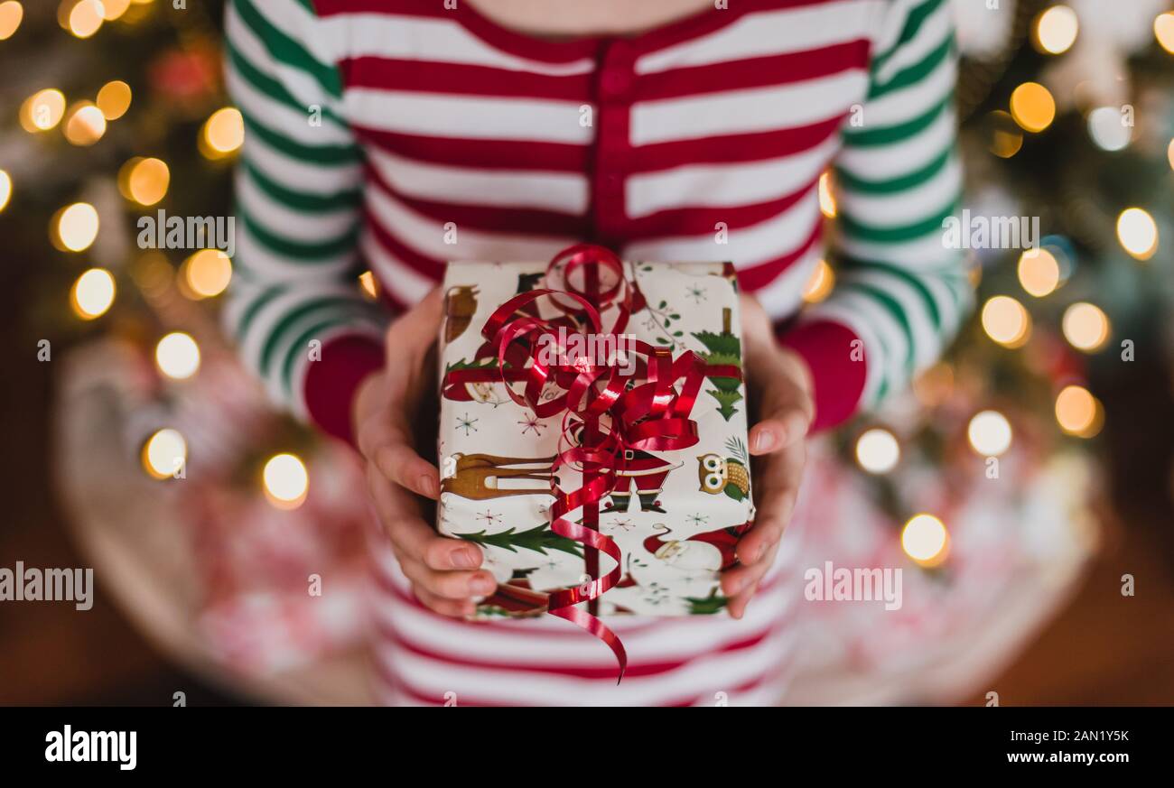 Bambino in pigiama che tiene regalo di Natale davanti alle luci sull'albero. Foto Stock