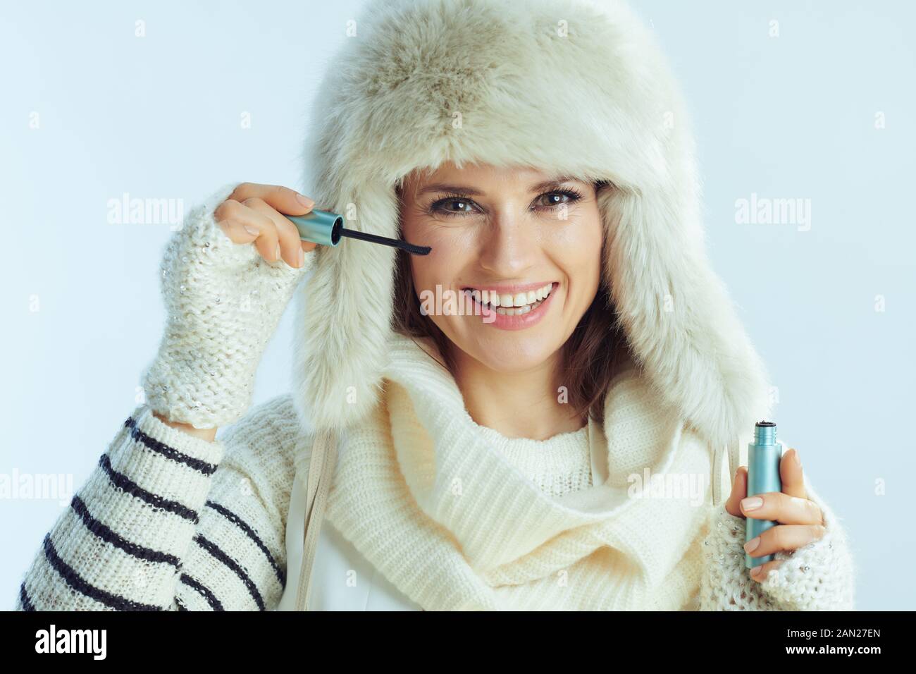 Ritratto di felice femmina elegante in bianco maglione a righe, sciarpa e cappello di lembi di orecchio utilizzando mascara contro inverno sfondo azzurro. Foto Stock