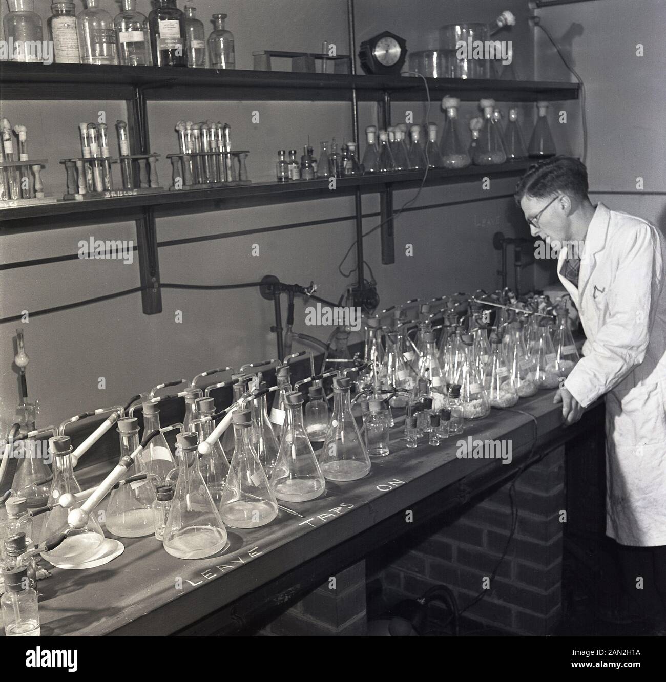 Degli anni Cinquanta, storico, maschio scienziato in un bianco-coat studiando una collezione di provette in vetro in un laboratorio, Università di Leeds, con le parole "Lasciare i rubinetti sull'' scritto sul banco di lavoro. Foto Stock
