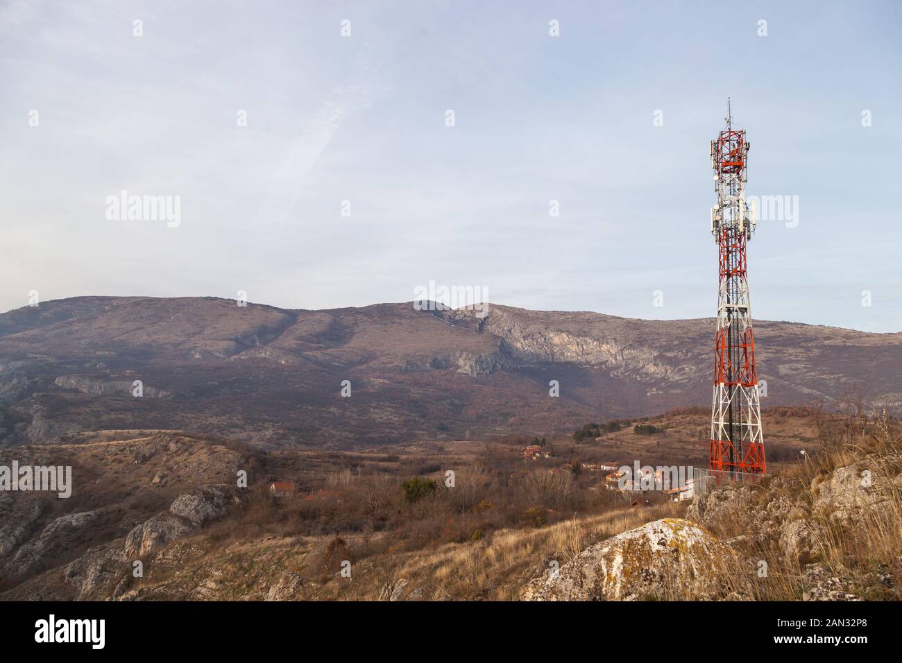 Torre di comunicazione rossa e bianca su una collina sopra il piccolo villaggio, cime rocciose distanti e cielo azzurro nebuloso Foto Stock