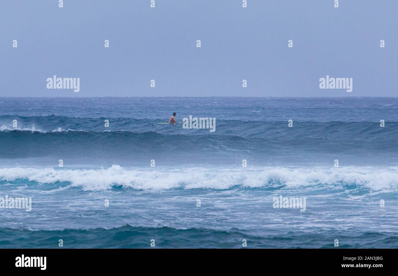 Lone surfer che guarda al mare come egli attende onde in condizioni di terreno accidentato Foto Stock