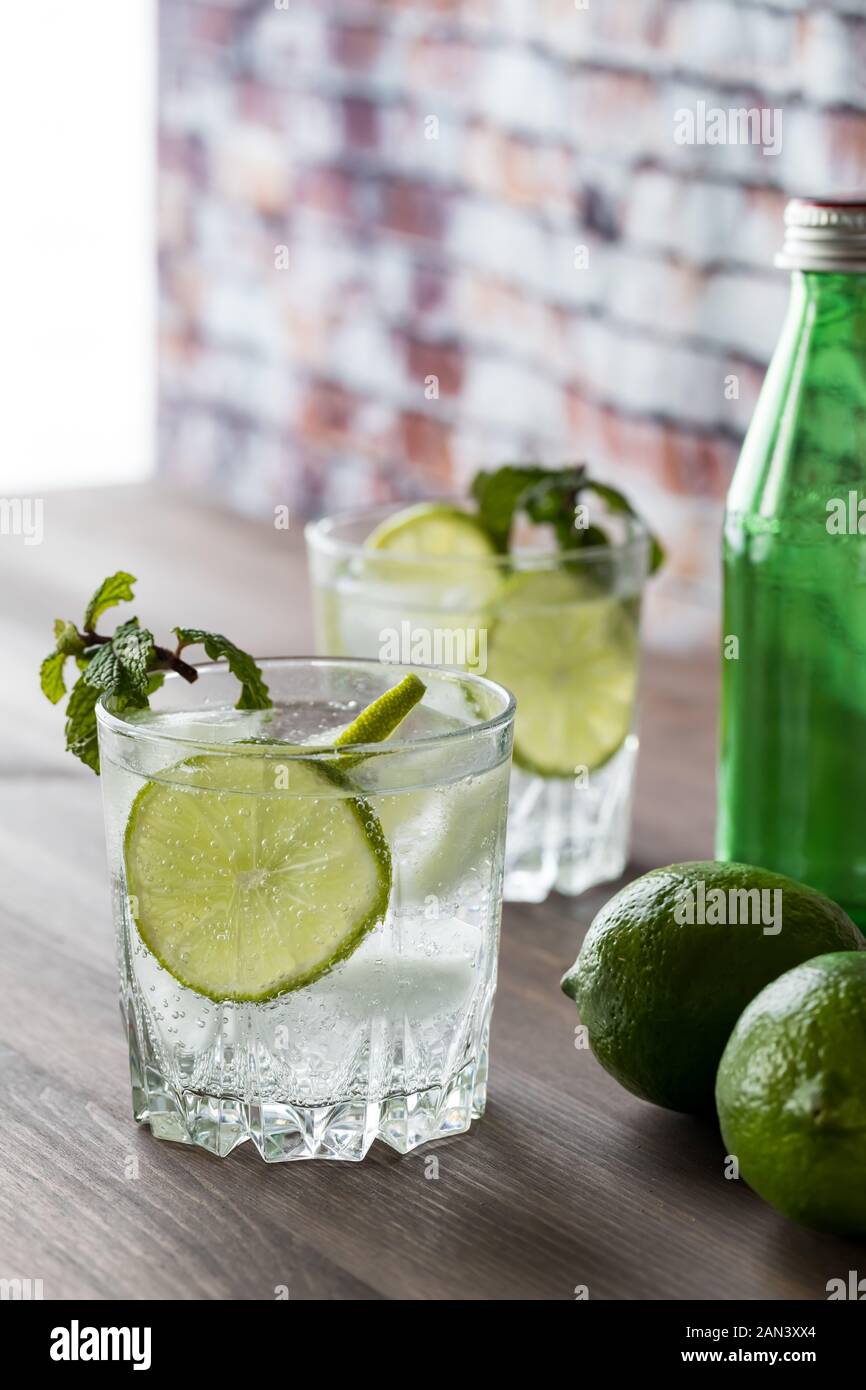 Un bicchiere di acqua frizzante con fettine di lime e menta su una tavola di legno contro una finestra piena di sole e di un muro di mattoni. Foto Stock