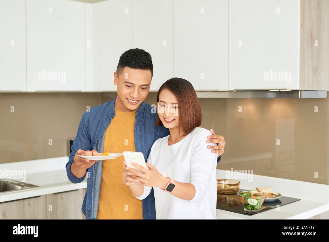 Romantico giovane coppia asiatica cucina insieme e utilizzando il telefono cellulare per selfie in bianco la cucina moderna, smart home concetto Foto Stock