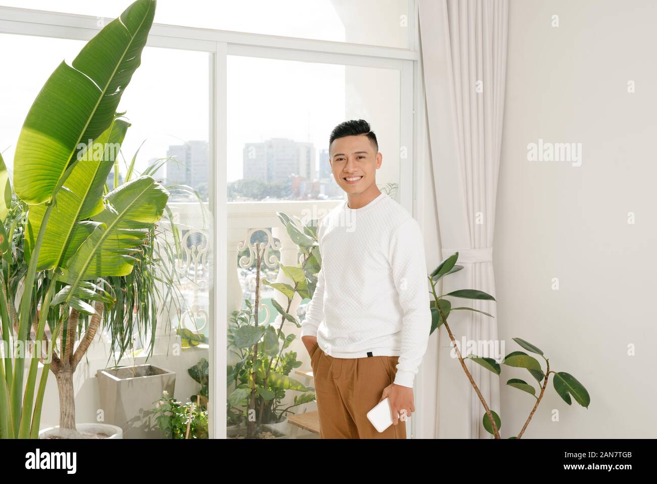 Asian giovane uomo sorridente nel soggiorno guardare fotocamera con tenere uno smartphone Foto Stock