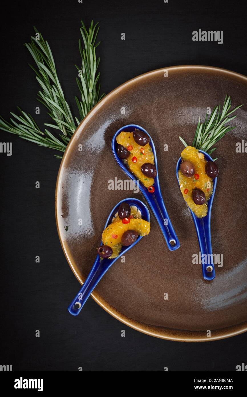 Vegano cibo sano. Crostino di Orange, oliva, peperoncino e rosmarino, servita su un cucchiaio di ceramica con uno sfondo nero. Foto Stock