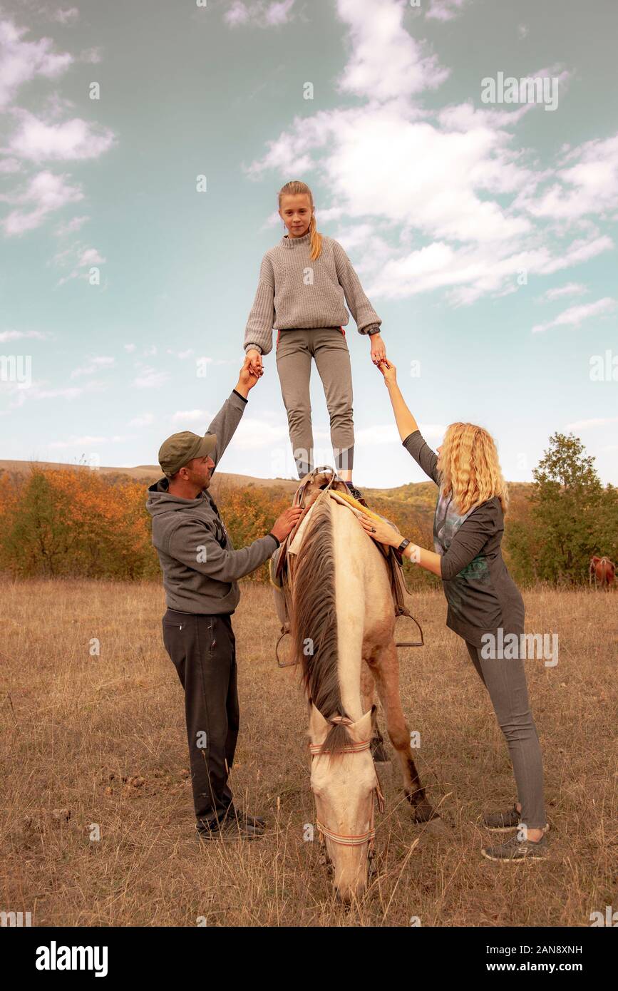 Una ragazza teenage si leva in piedi su un cavallo, sostenuto da entrambi i lati da adulti. Foto Stock