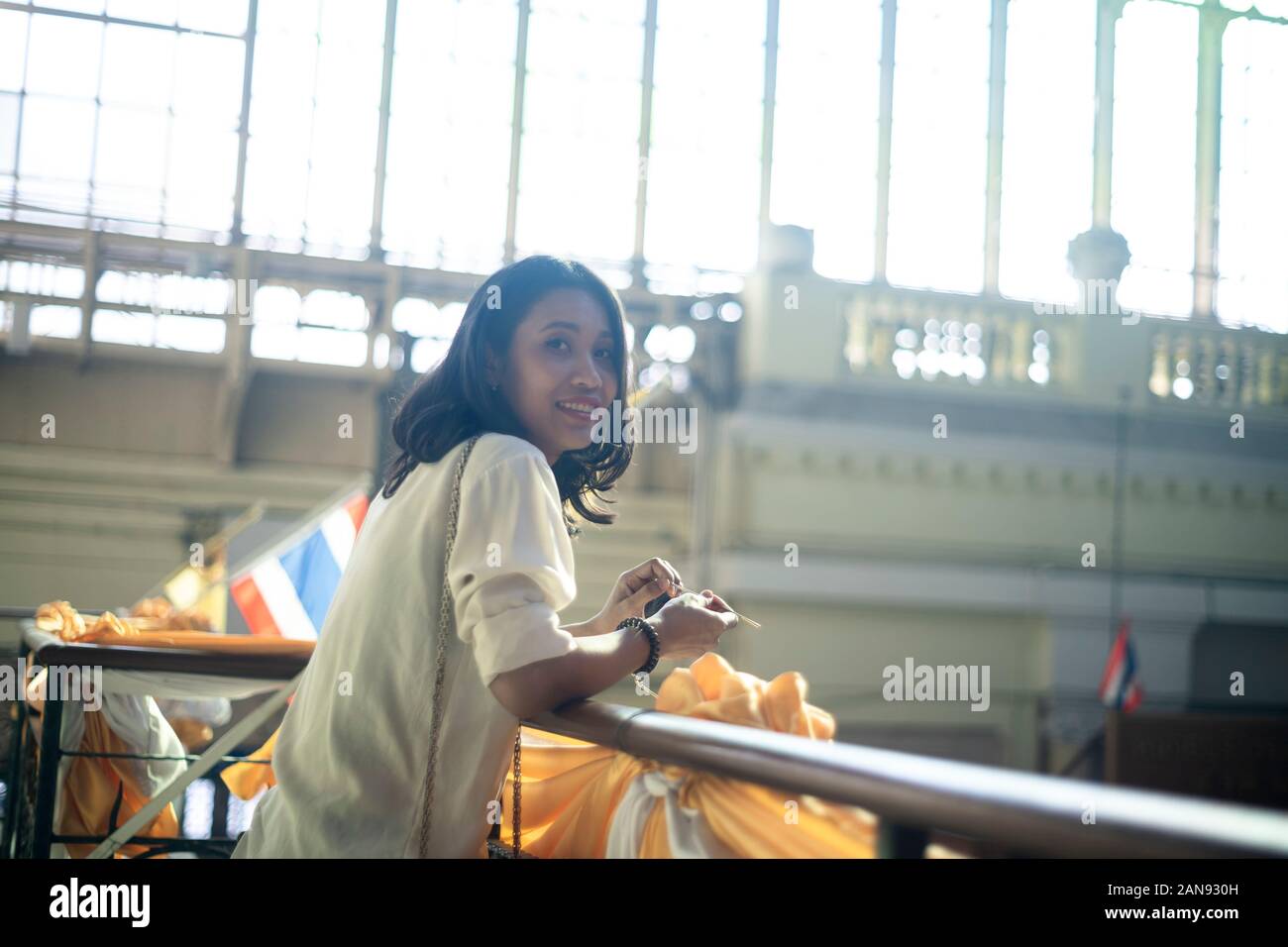 Il Nero dei capelli ondulati donna in maglia bianca permanente al corrimano di secondo piano nella stazione ferroviaria prop le braccia sul corrimano e sorriso per la fotocamera. Foto Stock