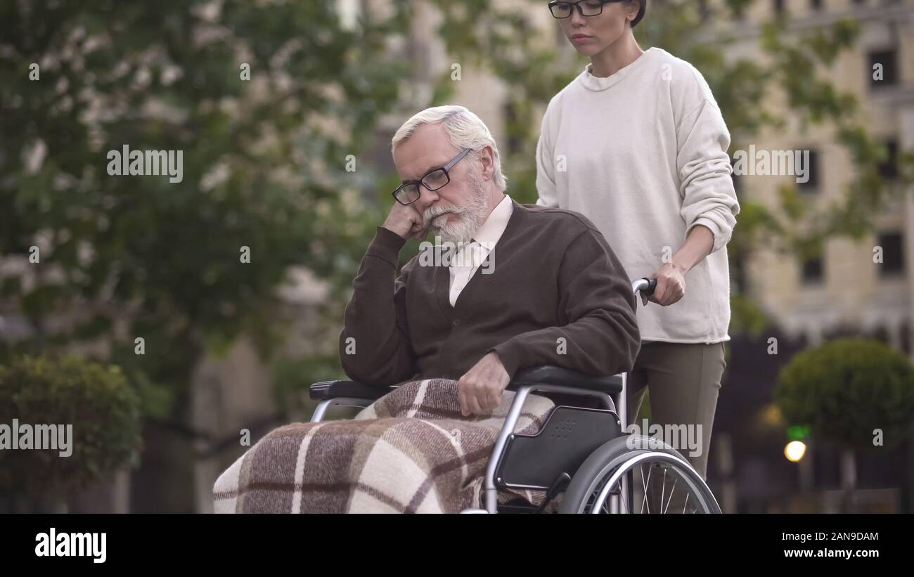 Giovane donna sulla passeggiata con triste pensionato in carrozzella, il caregiver assistenza, aiuto Foto Stock