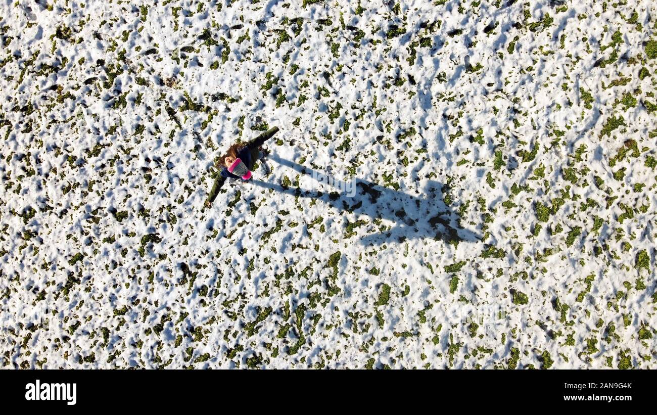 Vista aerea di una ragazza sul campo in erba ricoperta di neve in piedi e a diffondere il suo braccio e guardando alla propria ombra. Vista superiore da un drone Foto Stock