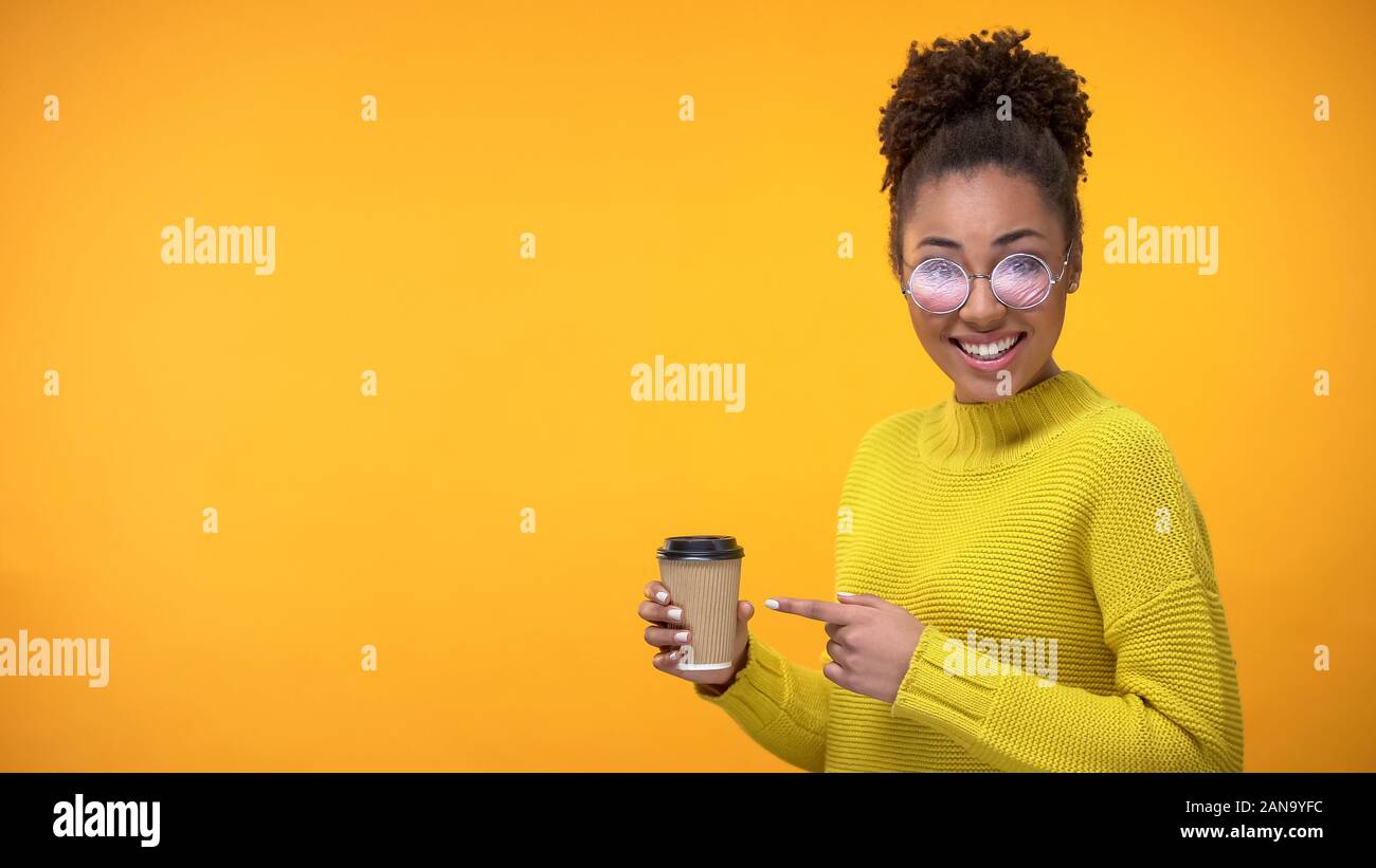 Elegante giovane donna puntando al bicchiere di carta di caffè e sorridente fotocamera, sorpresa Foto Stock