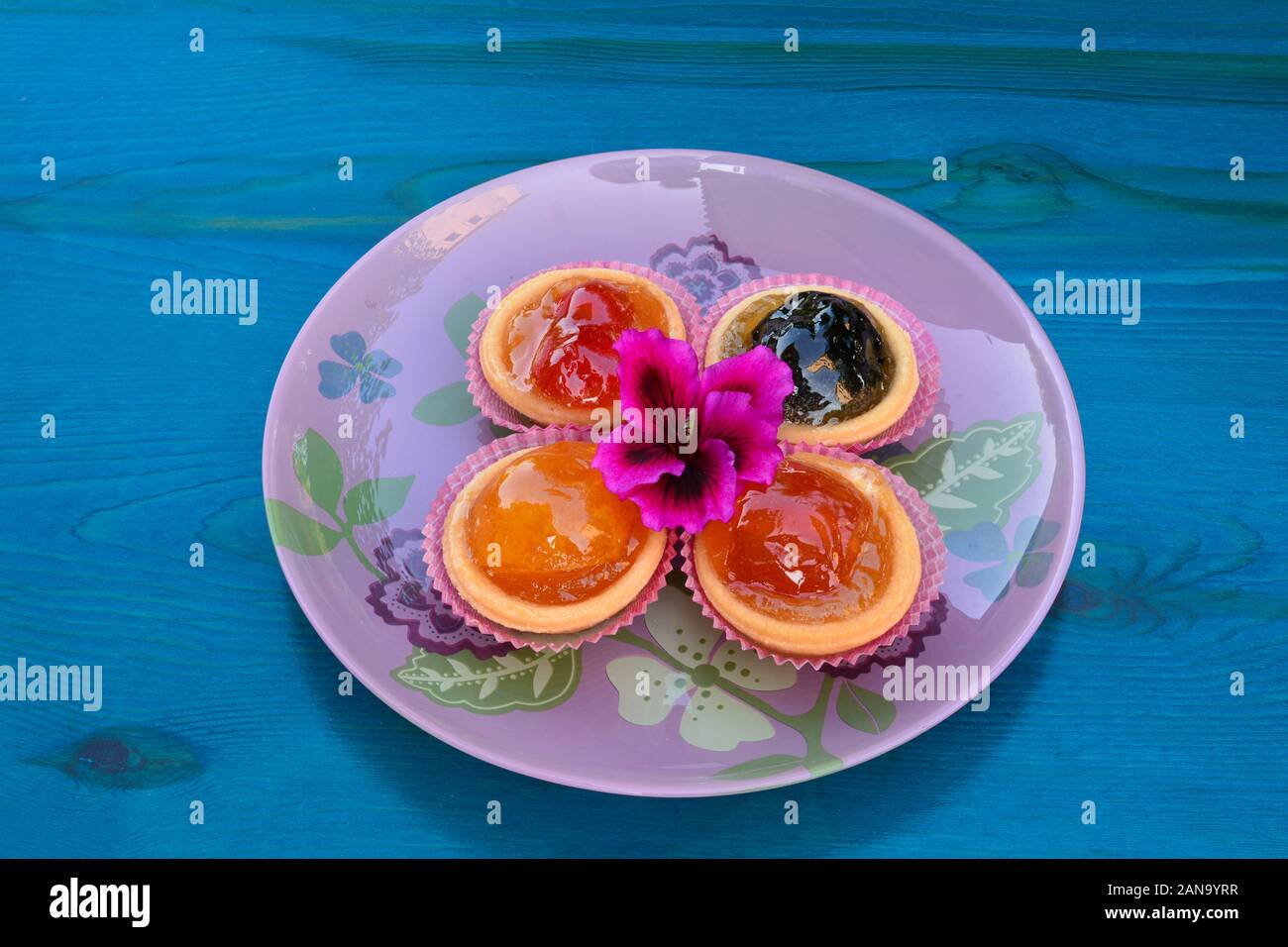 Punnets di pasta riempita con vari frutti canditi, servita in rosa lastra di vetro blu sul tavolo in legno e decorate con fiori di colore rosa Foto Stock