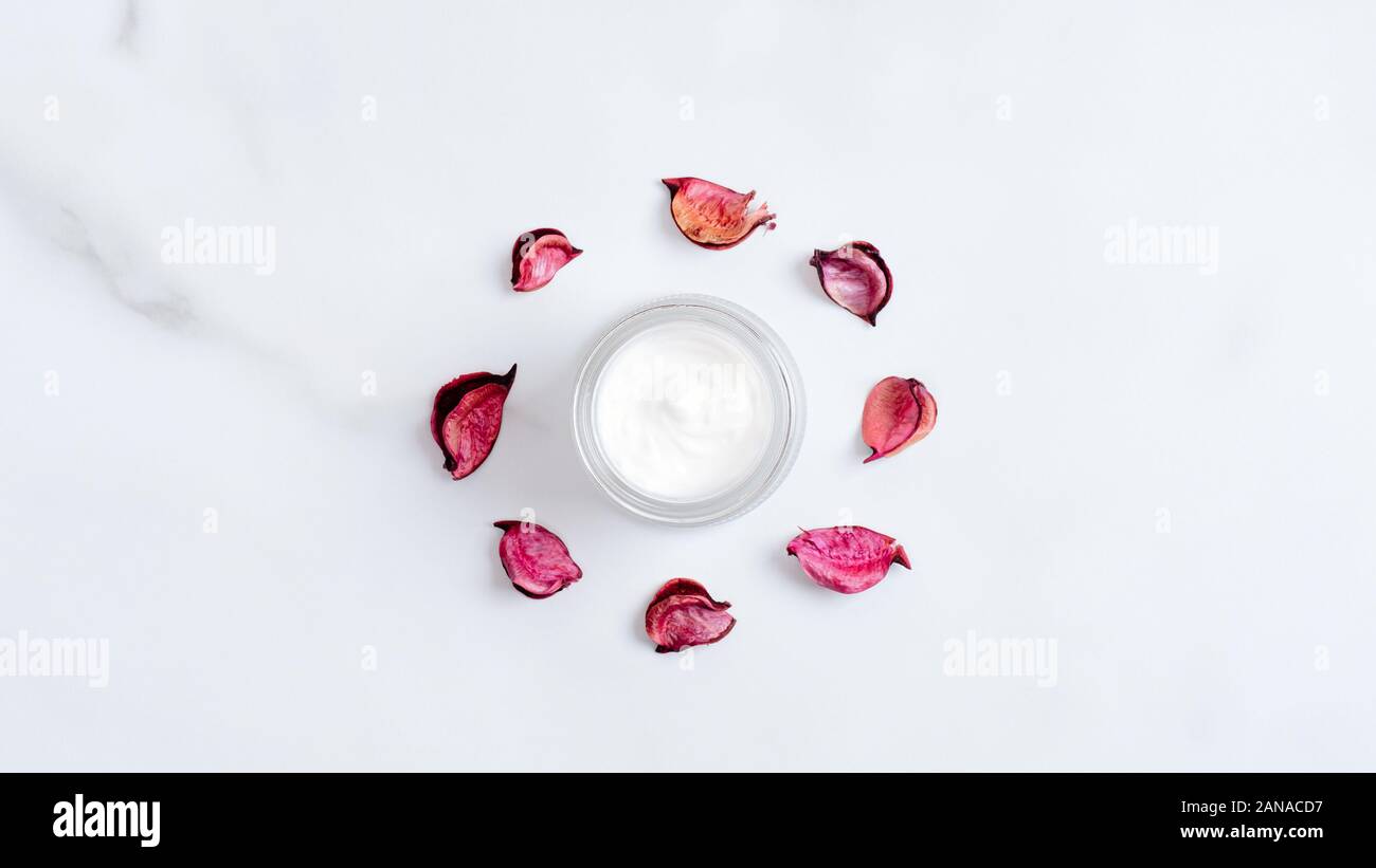 Organici di crema cosmetica jar rosso e petali di fiori su sfondo marmo. Vista superiore, piatto laici. Prodotti di bellezza naturali. Trattamenti viso e corpo il concetto di cura Foto Stock