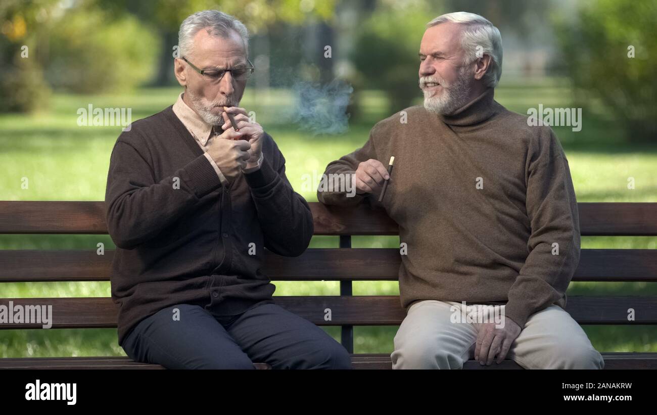 Due amici senior sigari di illuminazione, in appoggio su una panchina nel parco insieme, weekend Foto Stock