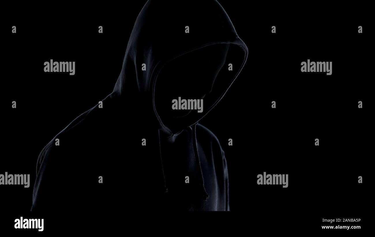 Pericoloso maniaco in maschera e hoodie in piedi contro lo sfondo nero, criminalità Foto Stock