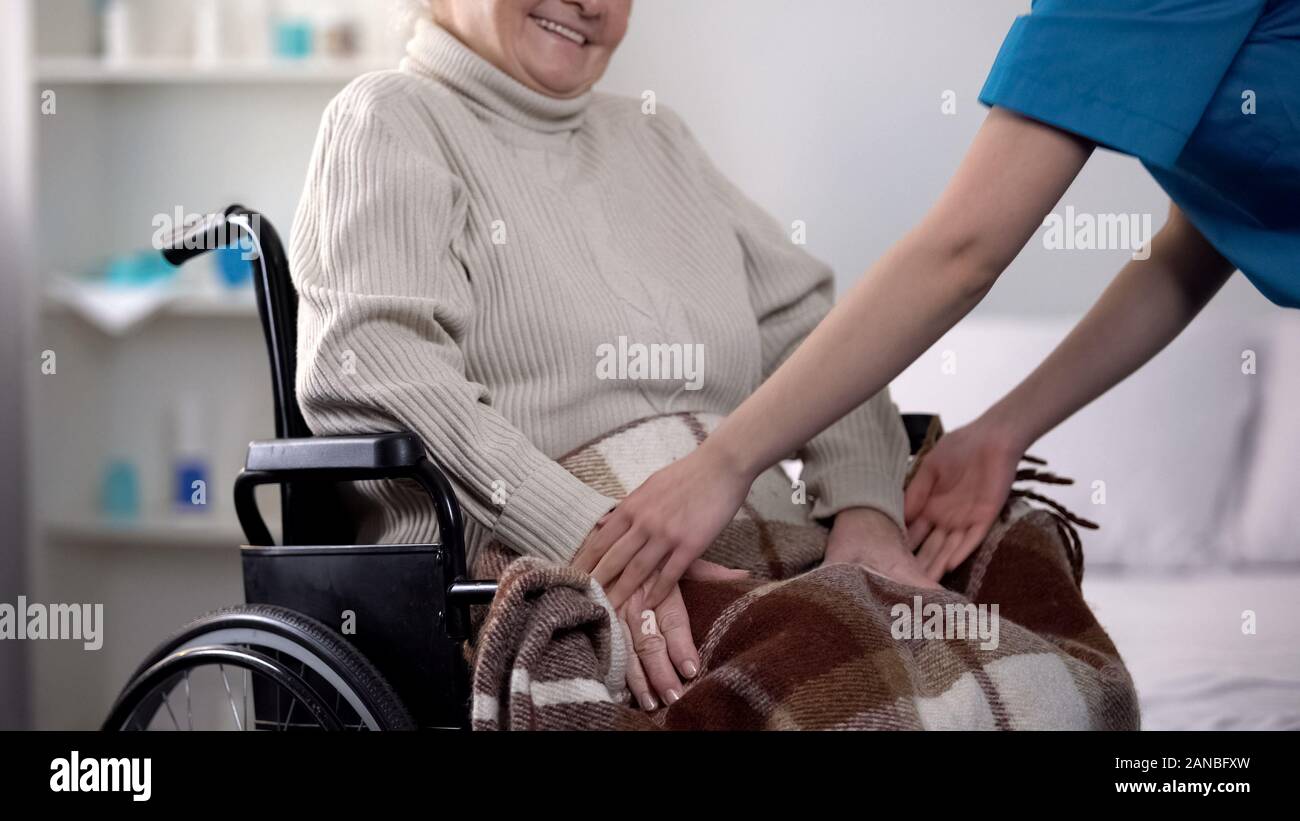 Infermiere coprendo delicatamente con coperta donna in carrozzella, cura circa le persone anziane Foto Stock
