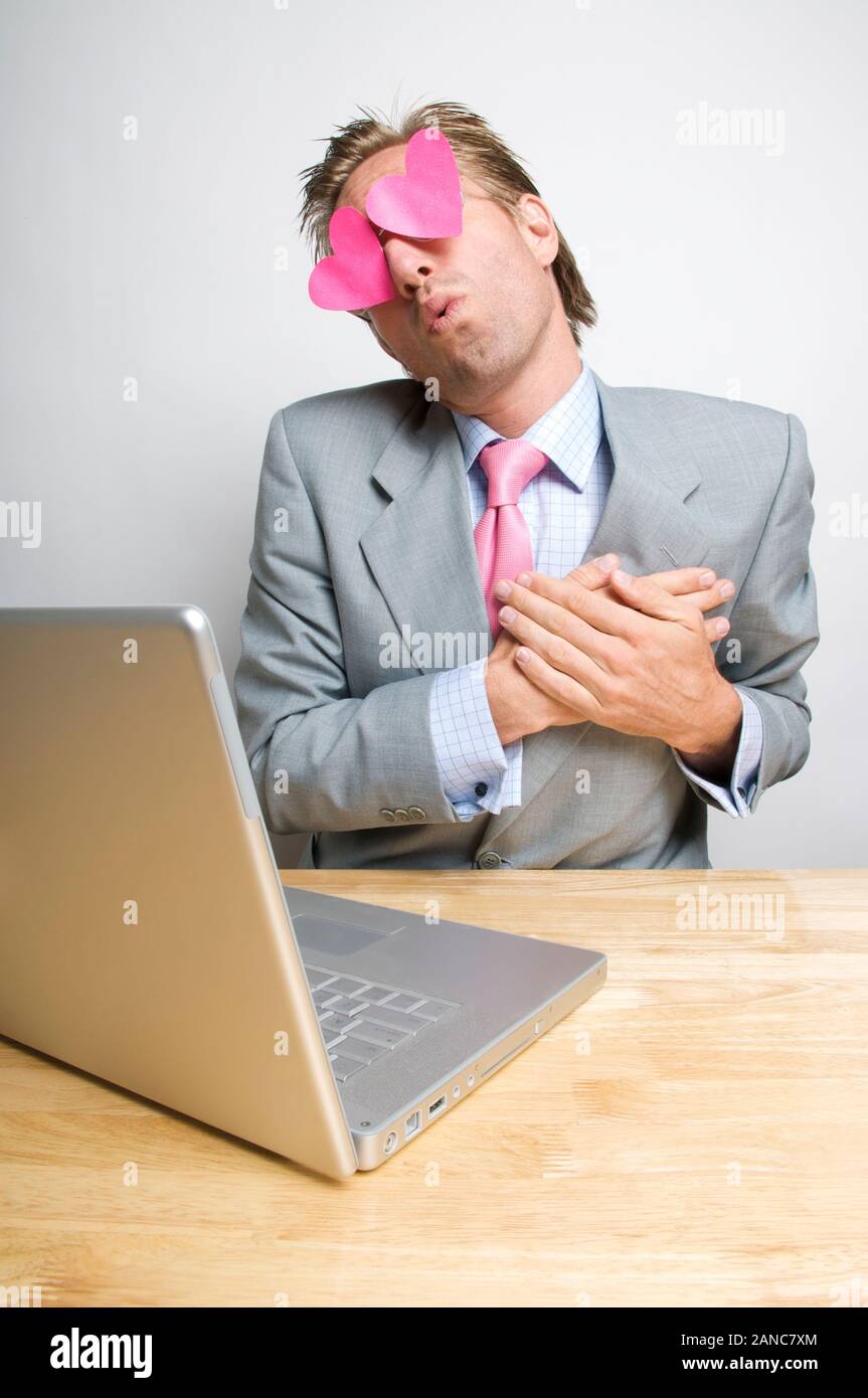 Imprenditore Lovestruck swooning seduto alla sua scrivania con il cuore ai suoi occhi guardando il computer portatile sulla sua scrivania Foto Stock