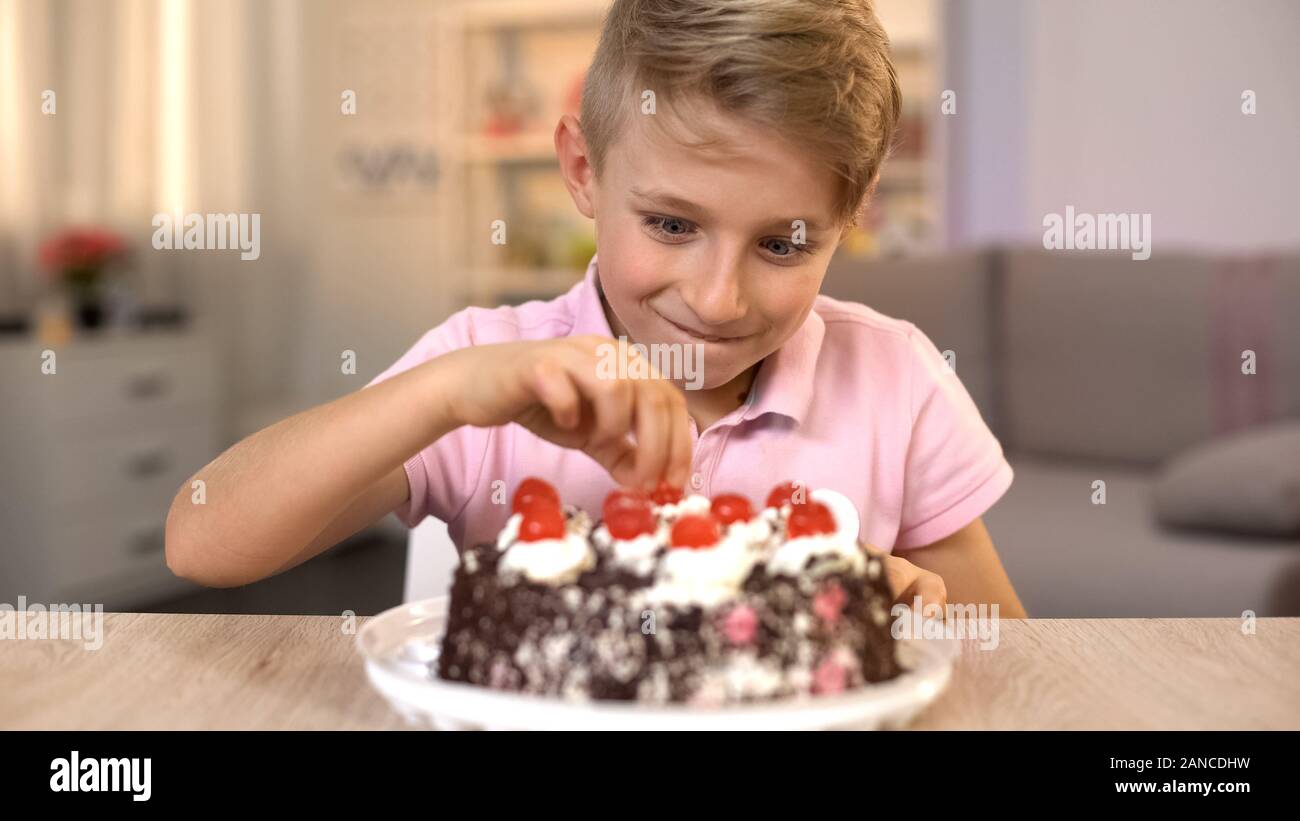 Ragazzo eccitato tenendo la ciliegia dalla parte superiore della torta al cioccolato, festa di compleanno Foto Stock
