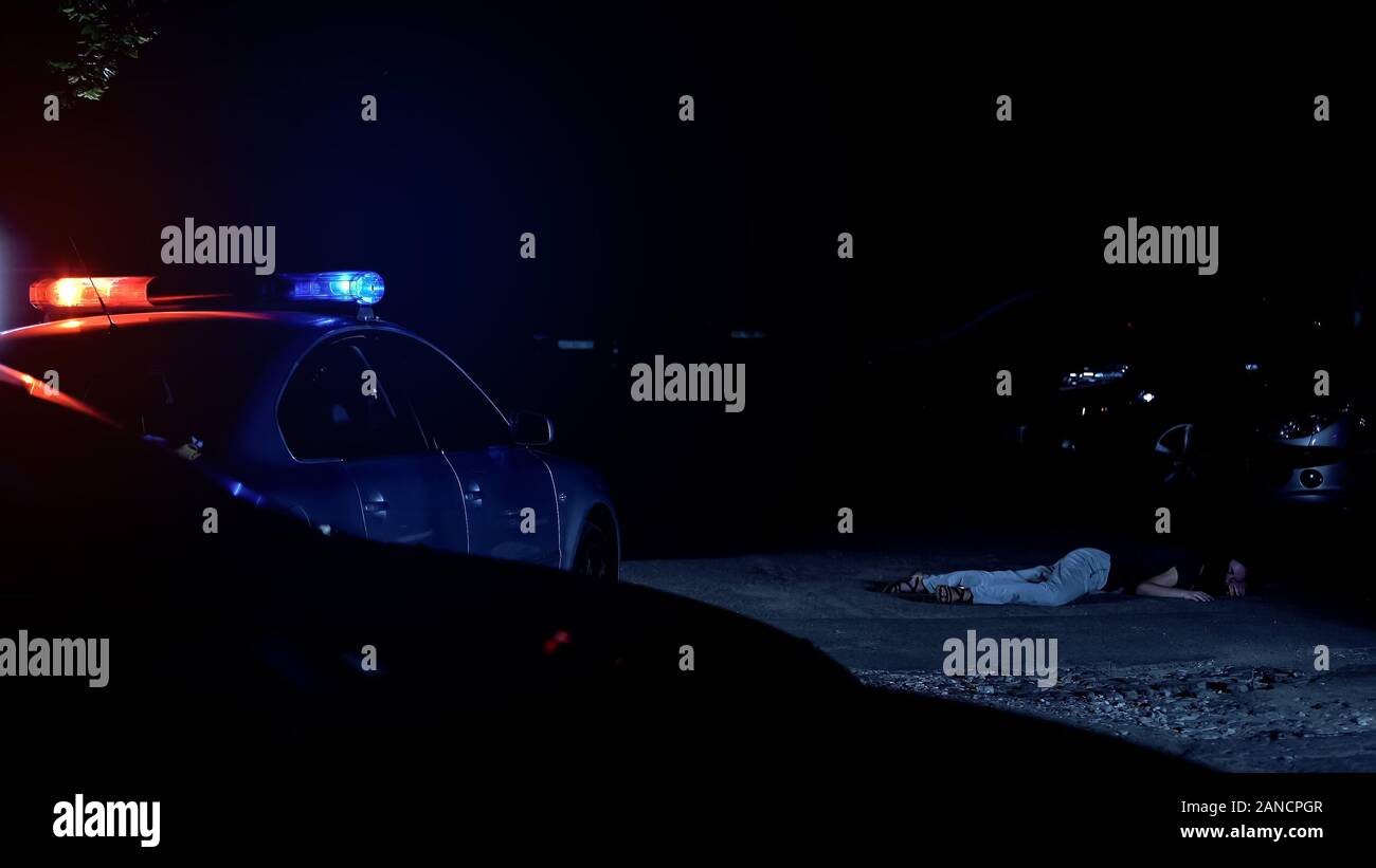 Auto di pattuglia con luci lampeggianti in piedi vicino alla vittima di un reato, giacente su strada Foto Stock