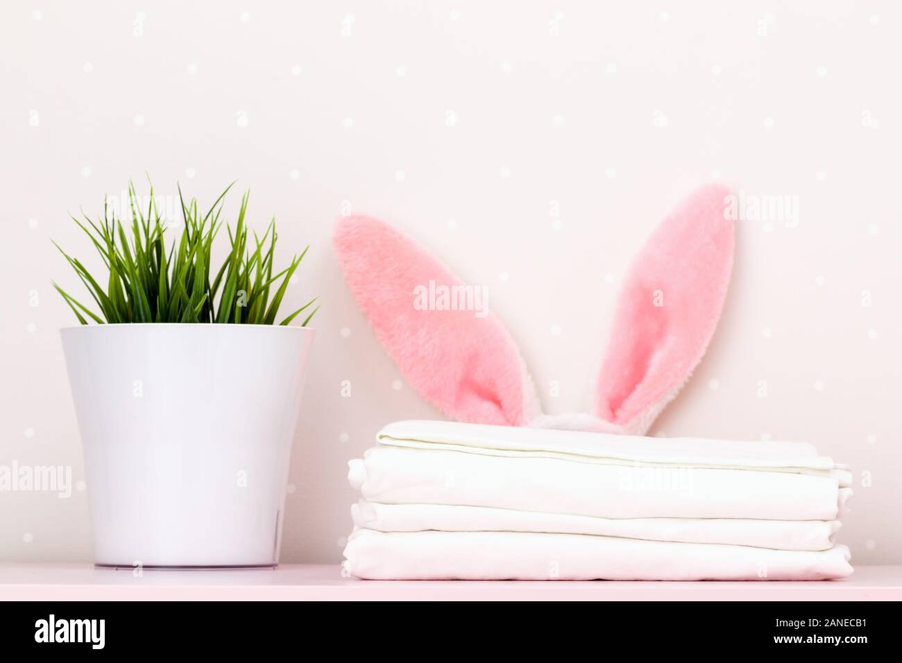 Un close-up di una pila di pulire biancheria da letto bianca, coniglietto pasquale per le orecchie e un houseplant, su un tavolo, contro uno sfondo di pareti di luce. Foto Stock