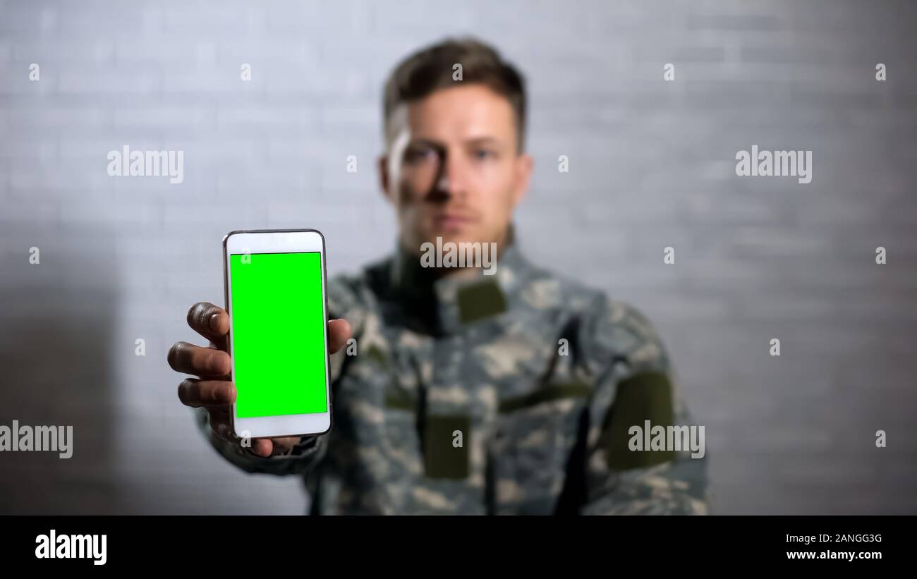 Soldato maschio in uniforme che mostra smartphone con schermo verde in telecamera, app Foto Stock