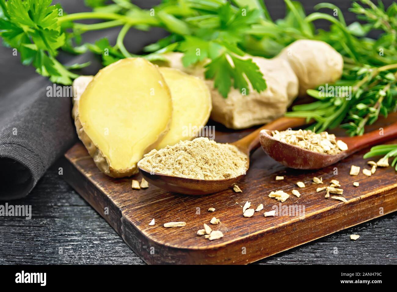 Lo zenzero in polvere e scaglie in due cucchiai di legno, radice di zenzero, erbe aromatiche, tovagliolo su una tavola di legno sfondo Foto Stock