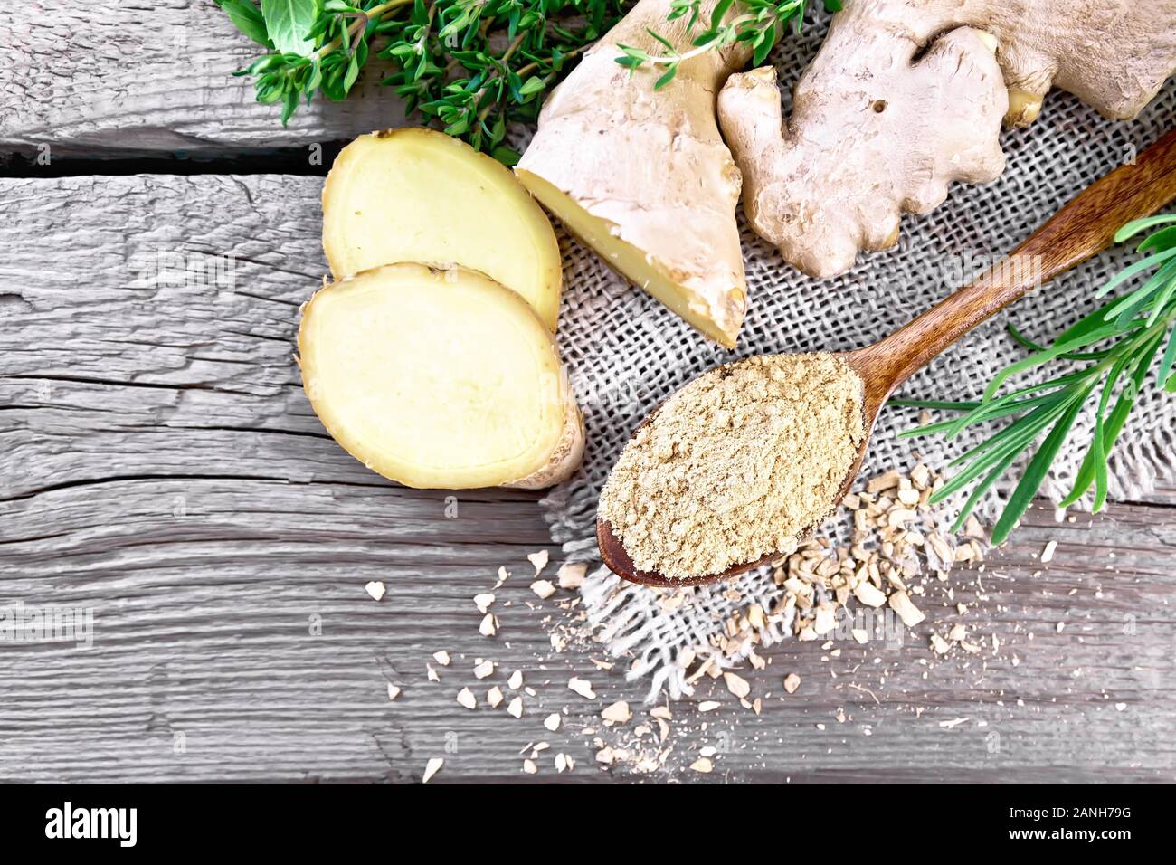 Lo zenzero in un cucchiaio, fiocchi e radice di zenzero su tela, erbe aromatiche su una tavola di legno del fondo da sopra Foto Stock