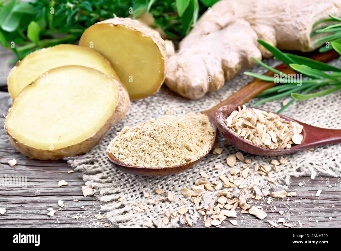 Lo zenzero in polvere e scaglie in due cucchiai di legno, radice di zenzero su tela, erbe aromatiche su uno sfondo di un vecchio pannello di legno Foto Stock