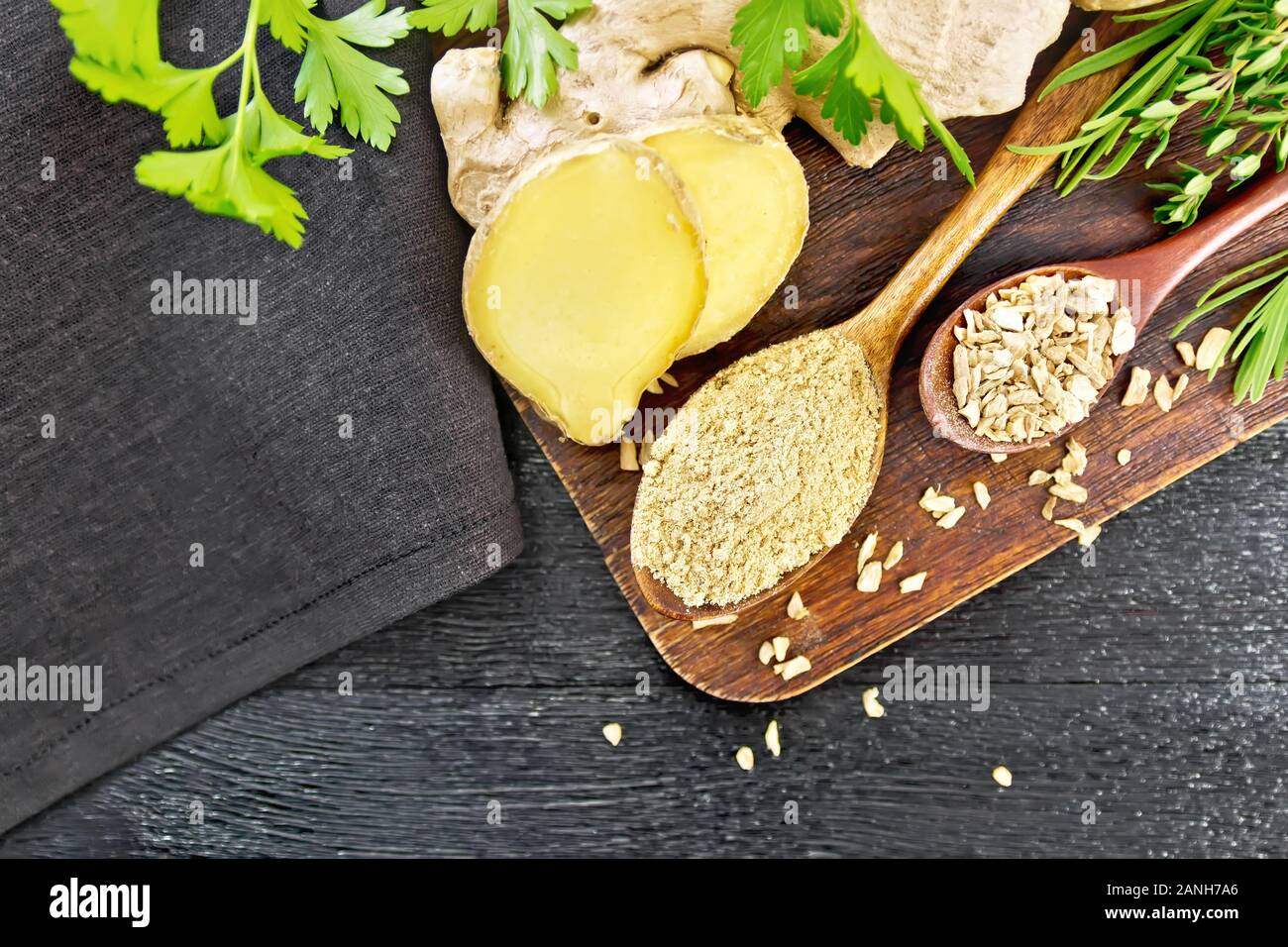 Lo zenzero in polvere e scaglie in due cucchiai di legno, radice di zenzero, erbe aromatiche, tovagliolo su una tavola di legno del fondo da sopra Foto Stock