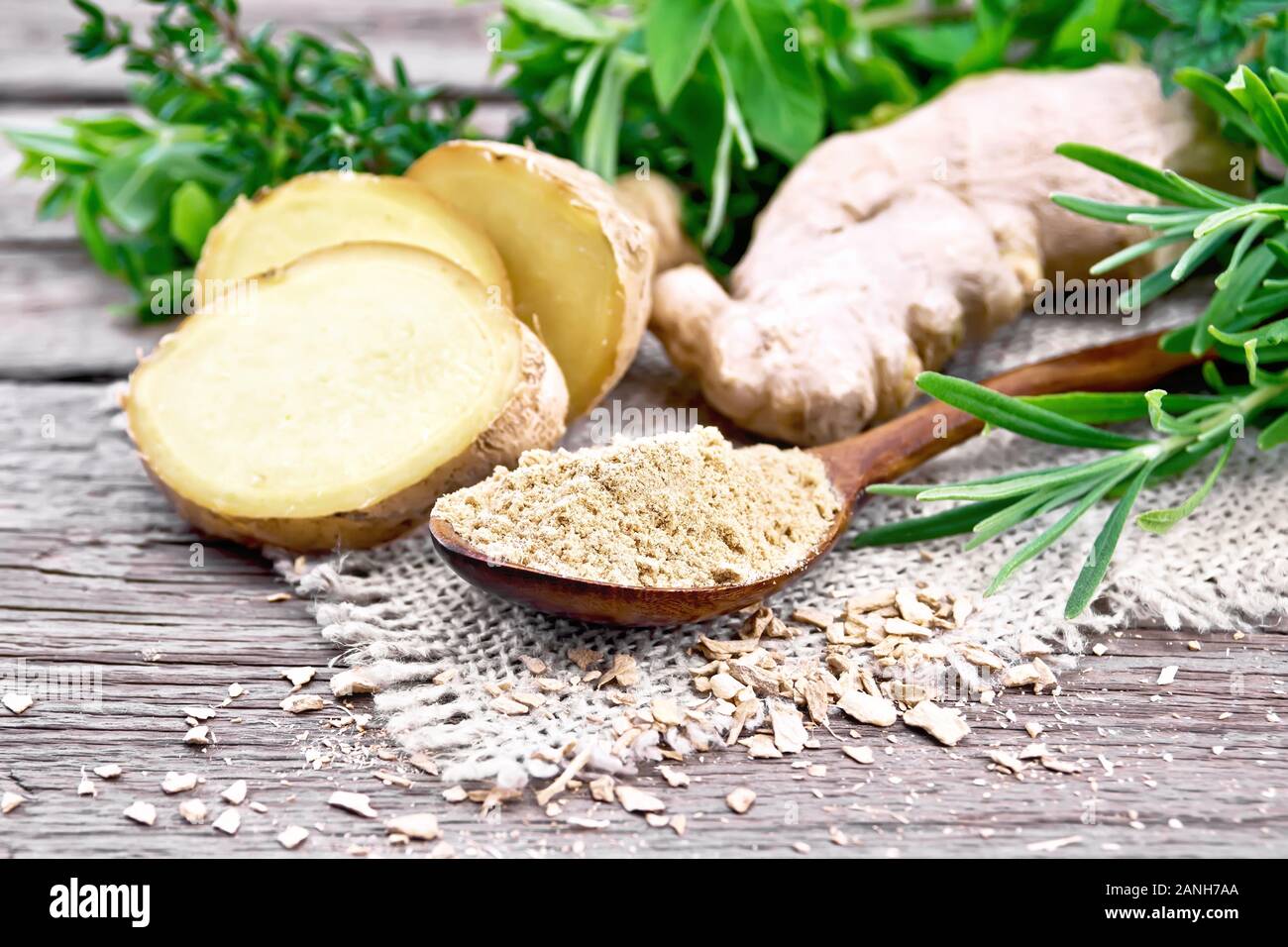 Lo zenzero in un cucchiaio, fiocchi e radice di zenzero su tela, erbe aromatiche su una tavola di legno sfondo Foto Stock