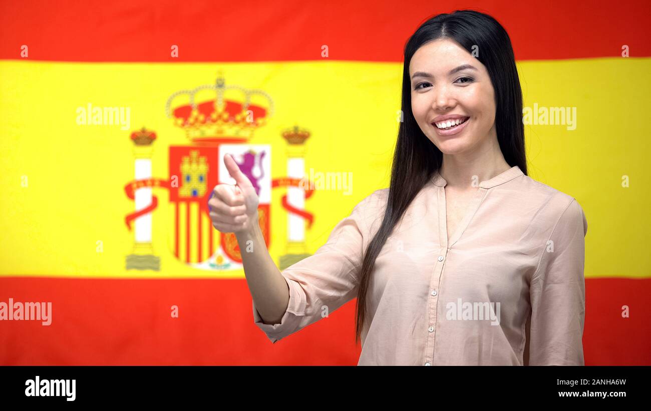 Sorridente donna asiatica che mostra Thumbs up gesto contro la bandiera spagnola sfondo Foto Stock